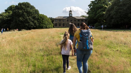 A mum and two children walk away from the camera towards the stables through the parkland along a mown path
